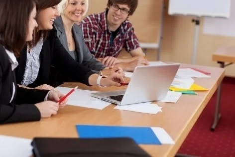 Un groupe d'étudiants souriants, collaborant autour d'une table lors d'un séminaire ou d'un atelier éducatif. Ils sont en train de discuter et de partager des idées, avec des documents, des ordinateurs portables et des fournitures de bureau devant eux. L'ambiance est celle d'un environnement d'apprentissage dynamique et interactif, typique des institutions éducatives au Québec, qui favorise l'échange culturel et l'engagement académique.