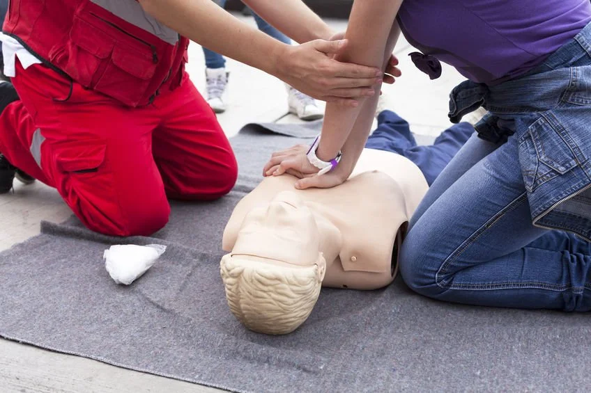 Des personnes en formation aux premiers secours pratiquent la réanimation cardio-pulmonaire (RCP) sur un mannequin. Un instructeur en combinaison rouge guide les mains d'un participant pour effectuer correctement les compressions thoraciques. Cela illustre un cours de formation pratique, une collaboration éducative entre la France et le Canada pour améliorer les compétences en matière de santé et de sécurité.