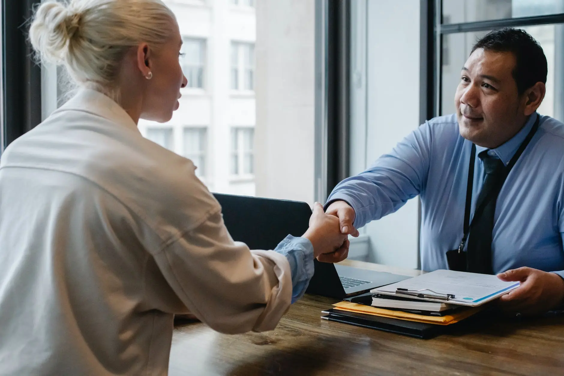 Photographie d'un entretien professionnel où une femme d'affaires et un homme d'affaires se serrent la main à travers une table de bureau, symbolisant une entente ou une embauche réussie. L'homme est assis et souriant, montrant une attitude accueillante, tandis que la femme, vue de dos, semble engagée dans l'action de serrer la main.